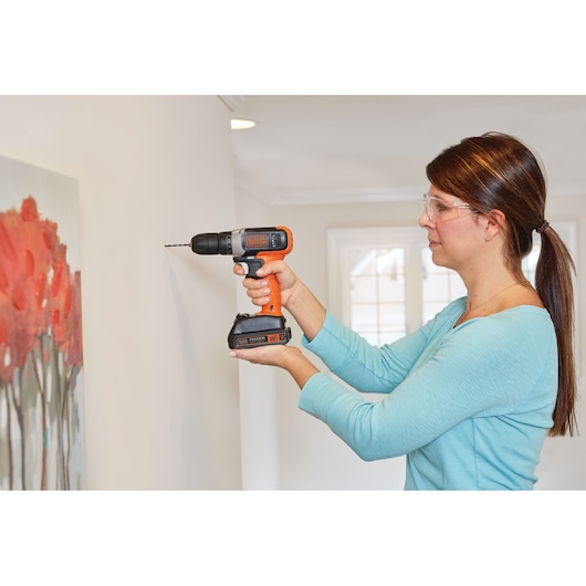 Person using BLACKDECKER BCD702C1 cordless drill on a white wall indoors, wearing a light blue shirt.