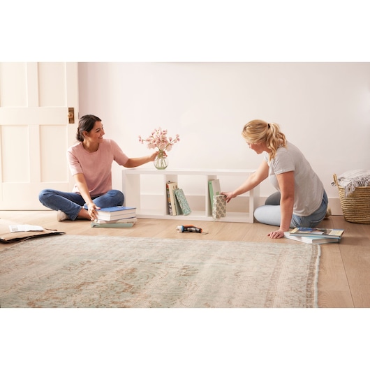 BLACKDECKER BCRTA601I cordless screwdriver on floor near two people organizing books and decor on white shelf.