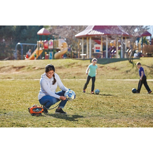 Photo of BLACKDECKER BDINF20 inflator used by a person to inflate a soccer ball on a field, with children playing nearby.