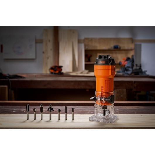 A BLACK+DECKER router tool in orange color is placed on a wooden workbench, with seven different router bits lined up in front of it. The background shows a workshop setting with tools and wood panels.