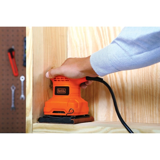 BLACKDECKER BS200 orange sander held by hand on wooden surface, workshop tools visible in background.