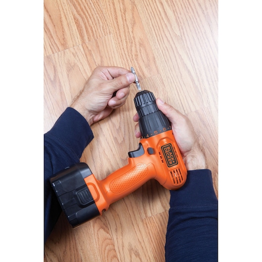 BLACKDECKER CD121K cordless drill in orange, shown from above on wood floor, person inserting drill bit.