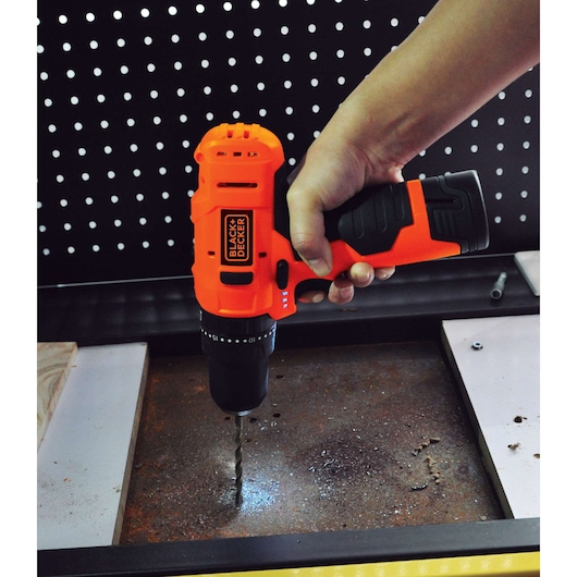 Photo of BLACK+DECKER HP12 cordless drill in use, drilling into a metal surface, black and orange design.