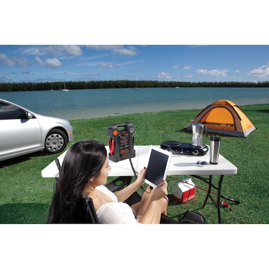 BLACKDECKER JS350S portable jump starter on outdoor table with camping gear, tent, and lake in background.