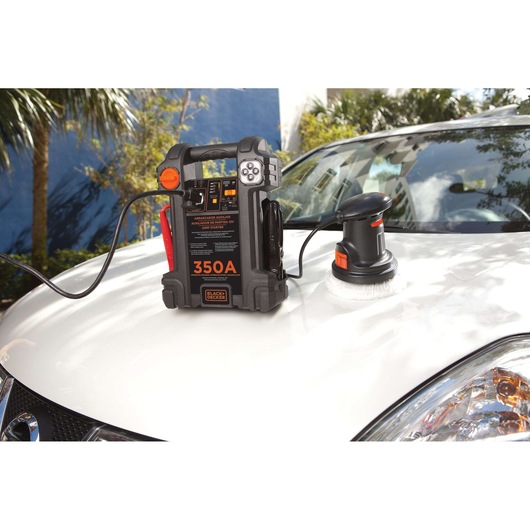 BLACK+DECKER 350A portable jump starter and a polisher on the hood of a white car, outdoors with trees and buildings reflected on the car’s surface.