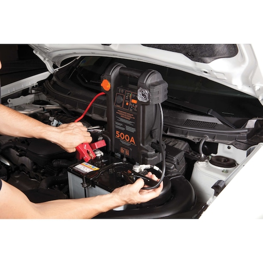 BLACKDECKER JS500S portable jump starter in use, connected to a car battery, hands attaching jumper clamps.