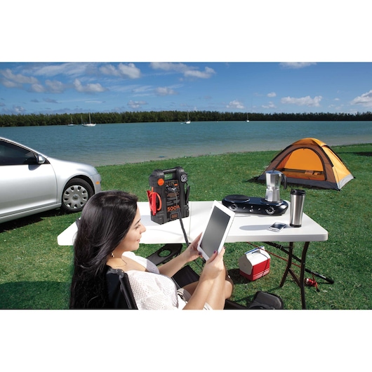 BLACKDECKER JS500S portable jump starter on a white table outdoors with camping gear, tent, and lake in background.